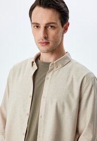 Camisa beige de botones hecha de tela suave, con un cuello clásico y una textura sutil, superpuesta a una camiseta de cuello redondo verde claro.