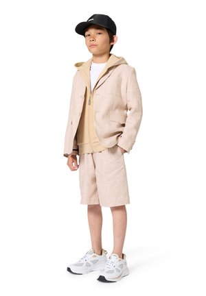 Blazer en lin beige porté sur un sweat à capuche marron clair, associé à un short beige et des baskets blanches. La casquette est noire avec un logo. Design propre et moderne.