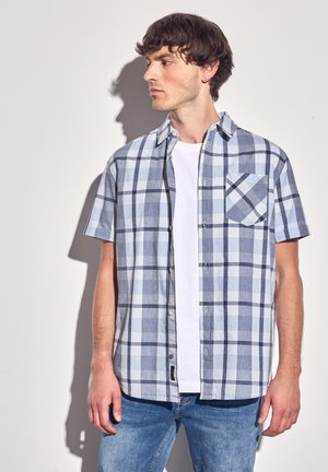 Camisa de botones de manga corta en patrón de cuadros azul claro y marino, con un único bolsillo en el pecho y un corte relajado. Llevada sobre una camiseta blanca.