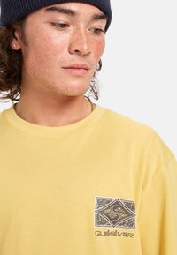Žluté bavlněné tričko s kulatým výstřihem, na levé části hrudi s grafickým designem zahrnujícím tvar diamantu a branding Quiksilver.