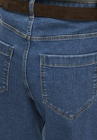 Jean en denim bleu avec une coupe classique, dotés de deux poches arrière, couture visible et texture lisse. Ceinture en daim marron à la taille.