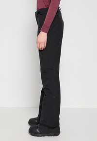 Zwarte waterdichte skibroek met een rechte pasvorm, verstelbare taille en versterkte stiksels, gecombineerd met zwarte geïsoleerde snowboots.