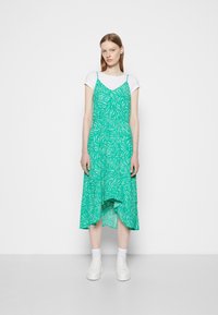 Vestido verde, com padrões, alças finas, decote em V e barra alta-lateral. Usado sobre uma t-shirt branca, combinado com ténis brancos.