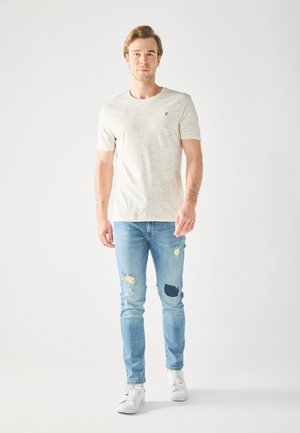 Lys beige T-shirt med kort ærme og en tekstureret overflade, forsynet med et lille teal-logo, parret med lyseblå jeans med slidte lapper.