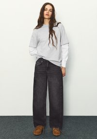 Žena s dlhými vlnitými vlasmi má na sebe vrstvené sivé a biele dlhé tričká, voľné čierne džínsy a hnedé mokasíny, stojí na koberci.