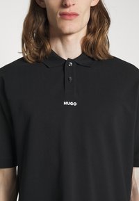 Tricou polo negru dintr-un material fin, cu guler. Dispune de o închidere cu butoane și un logo alb "HUGO" pe piept.