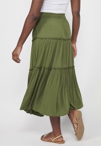 Femme portant une jupe fluide olive à volants et des sandales plates dorées, vue de dos sur un fond clair uni.