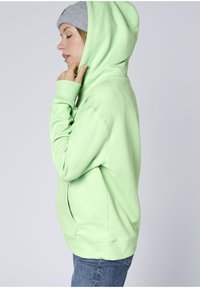 Hellgrüner Kapuzenpullover mit Kordelzug und Fronttasche, aus weichem Stoff gefertigt. Wird mit einer grauen Mütze getragen und seitlich vor einem neutralen Hintergrund präsentiert.