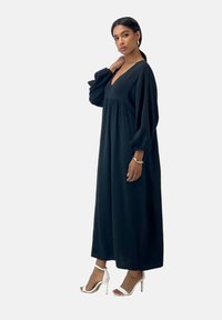 Lungo vestito nero con scollatura a V, maniche puff lunghe e corpino arricciato, abbinato a sandali bianchi con tacco alto. Tessuto liscio.