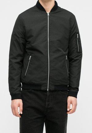 Personne portant une veste bomber noire avec des poches zippées sur le devant et la manche, associée à un pantalon noir, debout devant un fond blanc.