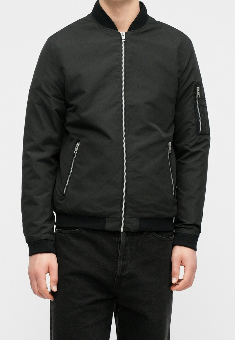 Personne portant une veste bomber noire avec des poches zippées sur le devant et la manche, associée à un pantalon noir, debout devant un fond blanc.