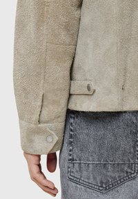 Beige mockaljacka med texturerad yta, med en nära knäppt ärm och ett tryckknapppar. Grå denimtyg synligt under.