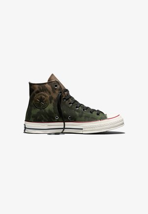 High-top Converse sneaker in groen en bruin camouflagepatroon met zwarte veters en een witte rubberen zool met rode en zwarte strepen.