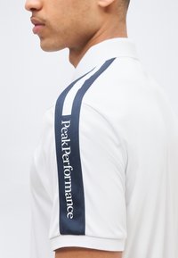 Gros plan sur une personne portant une chemise blanche avec une bande marine sur la manche, affichant le texte "Peak Performance" en lettres blanches.