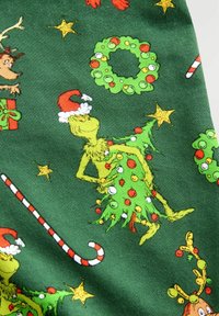 Grinch v Santa klobúku držiaci ozdobený vianočný stromček, obklopený venčekmi, cukrovými fontánkami, hviezdami a sobom na zelenom látkovom pozadí.