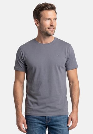 Uomo che indossa una t-shirt grigia semplice a maniche corte e jeans blu, che guarda leggermente verso destra su uno sfondo bianco.