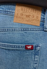 Hellblaue Jeans aus Denim mit einem hellbraunen Lederetikett mit geprägtem Text und einem roten Etikett mit einem Pferdelogo in der Nähe der Gesäßtasche.