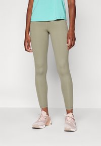 Leginsy w oliwkowym kolorze, wykonane z elastycznego materiału, o gładkim, dopasowanym kroju. Połączone z jasnoróżowymi butami sportowymi i turkusową bluzką.