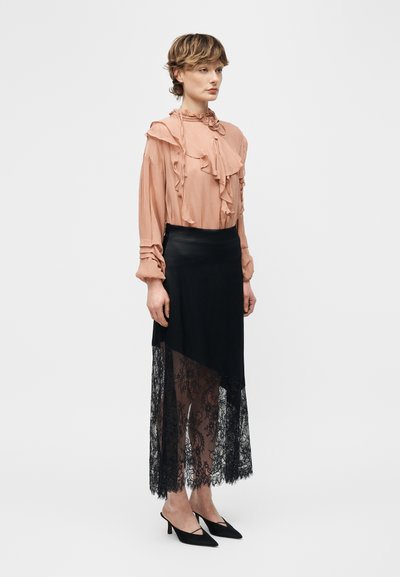 Γυναίκα με κοντά μαλλιά που φοράει διάφανη ροδακινί μπλούζα με βολάν, μαύρη φούστα με δαντέλα και μαύρα μυτερά τακούνια, στέκεται μπροστά σε λευκό φόντο.