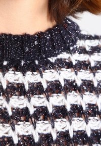 Haut tricoté présentant un motif à maille ajourée bleu marine et blanche, avec une finition texturée et des accents scintillants le long du décolleté.