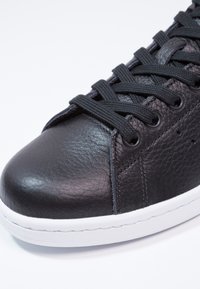 Černé kožené tenisky s texturovaným povrchem, kulatou špičkou, černými tkaničkami a bílou gumovou podrážkou. Obsahují perforované detaily na straně.