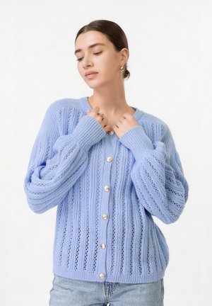 Kobieta ubrana w jasnoniebieski dziany kardigan z złotymi guzikami, trzymająca kołnierz, na jednolitym białym tle.