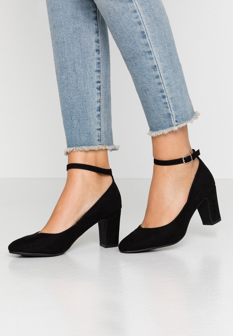 Chaussures en daim noir à talon moyen avec brides à la cheville portées avec un jean bleu clair à ourlet effiloché.