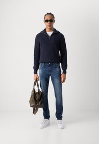 Marineblå strikket pullover med lynlåskrave, blå denim jeans, hvide sneakers og olivengrønt taske. Har ribbet tekstur og kontrasterende accenter.