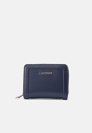 Cartera de cuero azul marino con una superficie texturizada, cierre de cremallera y logo plateado de Lacoste en la parte frontal. Diseño rectangular compacto.