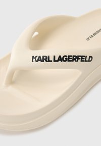 Béžové žabky vyrobené z gumy s jemne textúrovanou podrážkou. Obsahujú dizajn na medzičlánku a výrazné čierne logo "KARL LAGERFELD" na vrchnej časti.