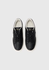 Pár černých kožených tenisek Tommy Hilfiger s bílými podrážkami a černými tkaničkami, zobrazených zeshora na bílém pozadí.