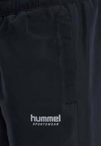 Čierne športové nohavice vyrobené z hladkého materiálu, s bočnou kapsou a strieborným logom "hummel SPORTSWEAR" blízko spodnej časti.