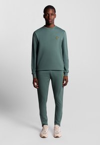 Groene sportieve trainingsoutfit van gladde stof, met een ronde hals, elastische manchetten en een klein geel logo op de borst.