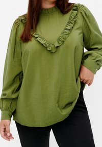 Kvinna som bär en olivgrön långärmad blus med volangdetalj i V-form och puffärmar, tillsammans med svarta byxor.