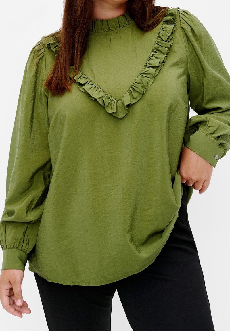 Kvinna som bär en olivgrön långärmad blus med volangdetalj i V-form och puffärmar, tillsammans med svarta byxor.