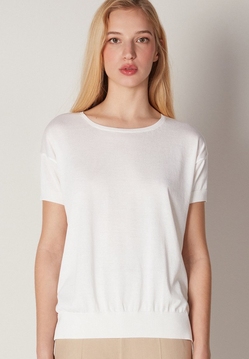 Biała krótka bluzka z krótkim rękawem, wykonana z lekkiego materiału, z okrągłym dekoltem i marszczonym dołem. Gładka tekstura, luźny krój, minimalistyczny design.