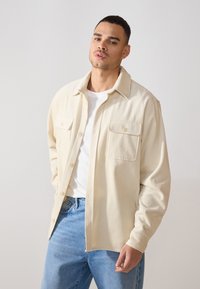 Chemise en velours côtelé beige avec un devant à boutons, deux poches poitrine et un col, portée sur un t-shirt blanc et un jean bleu.