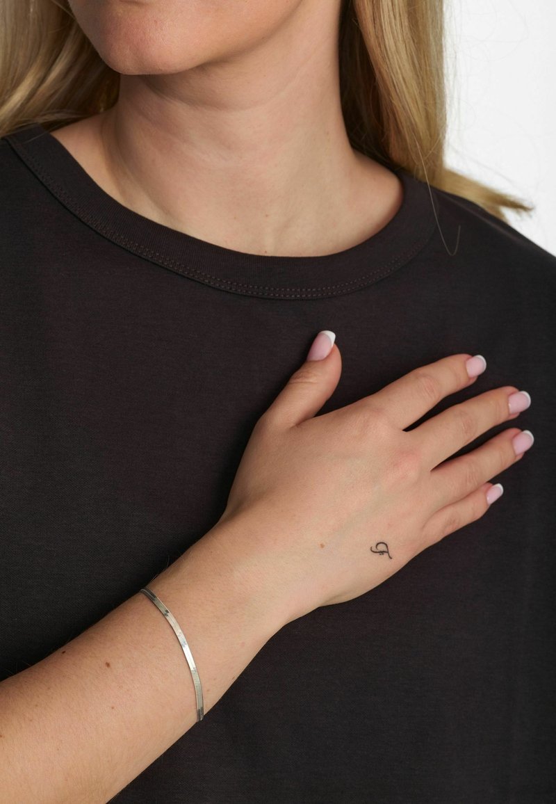 Chemise noire à manches courtes avec un col rond ; un bracelet manchette en métal argenté au poignet ; ongles manucurés rose clair ; petit tatouage sur la main.