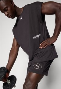 Svart ermeløs skjorte og shorts med hvite logoer; stoffet ser mykt ut. Personen løfter en vekt, som fremviser en muskuløs arm og detaljerte hardware.