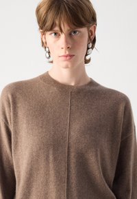 Καφέ πλεκτό πουλόβερ με στρογγυλή λαιμόκοψη, που διαθέτει αδιάκοπη σχεδίαση και μαλακή υφή, φορεμένο από ένα άτομο με κοντά μαλλιά και σκουλαρίκια.