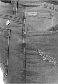 Jean en denim gris avec une zone usée sur la cuisse, des boutons métalliques et une poche arrière étiquetée avec un logo cousu.