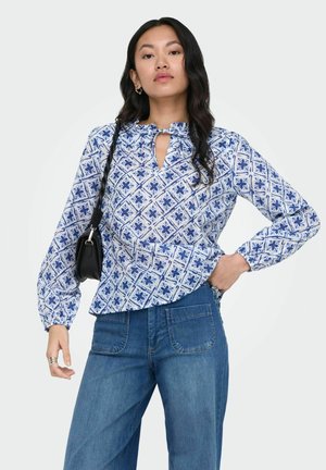 Femme aux longs cheveux noirs portant un chemisier bleu et blanc à motifs, un jean bleu et un sac à bandoulière noir, debout, une main sur la hanche.