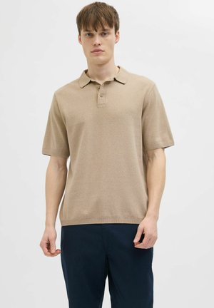 Junger Mann, der ein beiges kurzärmeliges Poloshirt und dunkelblaue Hose trägt, steht vor einem schlichten weißen Hintergrund.