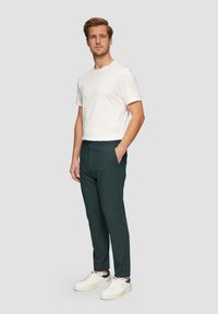 Weißes T-Shirt mit Rundhalsausschnitt, dunkelgrüne Hose mit glatter Textur und weiße Sneakers mit marineblauen Akzenten, stehend vor einem hellgrauen Hintergrund.