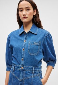 Πουκάμισο από τζιν με μυτερό γιακά, καστανά κουμπιά, τσέπη στο στήθος και φουσκωτά μανίκια. Το ύφασμα έχει μεσαίο μπλε χρώμα με υφή.