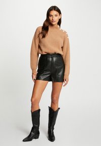 Pull en maille marron avec des boutons aux épaules, associé à une mini-jupe en cuir noir et des bottes de cowboy noires. Tenue décontractée avec des textures contrastées.