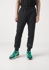 Pantaloni sportivi neri con vita regolabile, orli alle caviglie e tre strisce grigie nella parte inferiore delle gambe, abbinati a scarpe da ginnastica verdi.