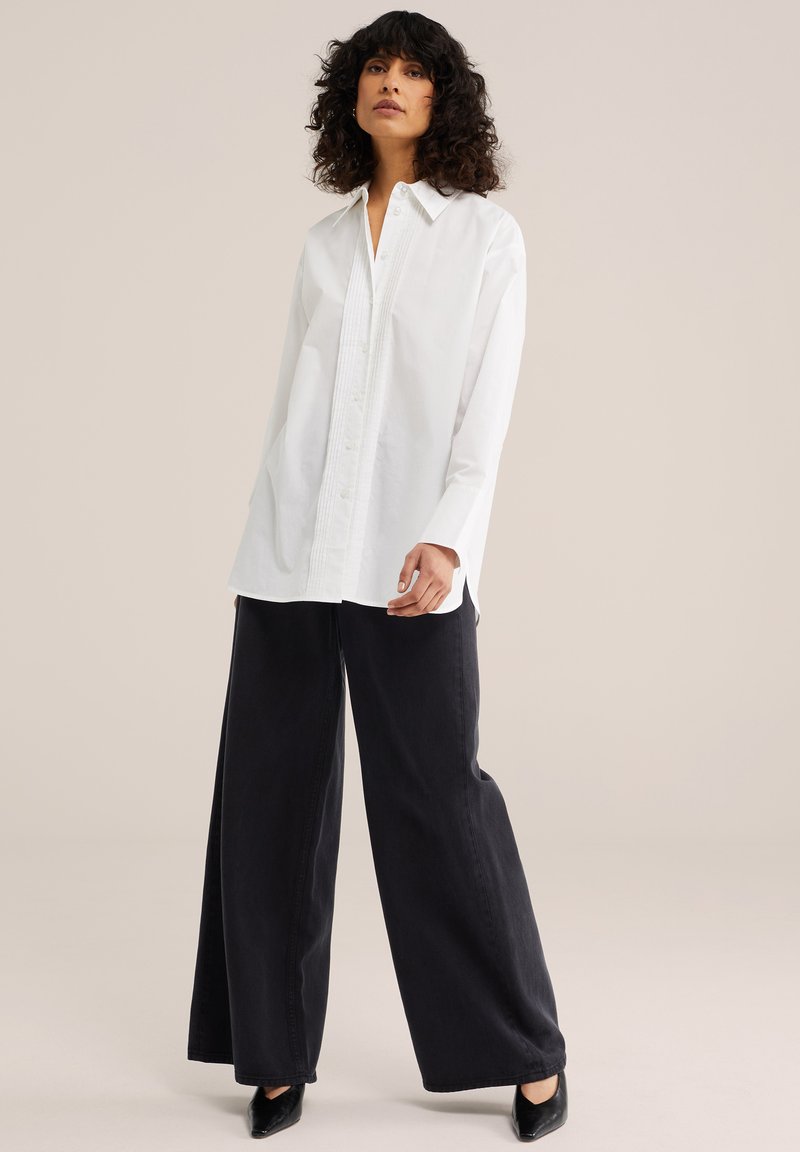 Femme aux cheveux bouclés portant une chemise blanche boutonnée, un pantalon ample noir et des chaussures noires à bout pointu, debout devant un fond neutre.