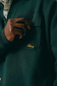 Mørkebrun hånd som justerer en gullring nær lommen på en mørkegrønn strikkegenser med en gul brodert logo.