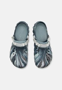 Chunky clogs made of navy and grey patterned rubber, featuring ventilation holes and a white adjustable strap on the heel.
